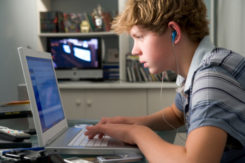 Young Boy In Bedroom Using Laptop And Listening To MP3 Player